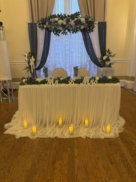 Mesa de novios con letrero Mr & Mrs, arco de flores azules y blancas, cortinas y velas